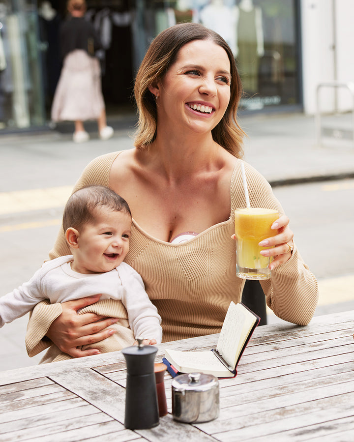 Momcozy M6 hands-free breast pump in use by smiling mother at a café with her baby.