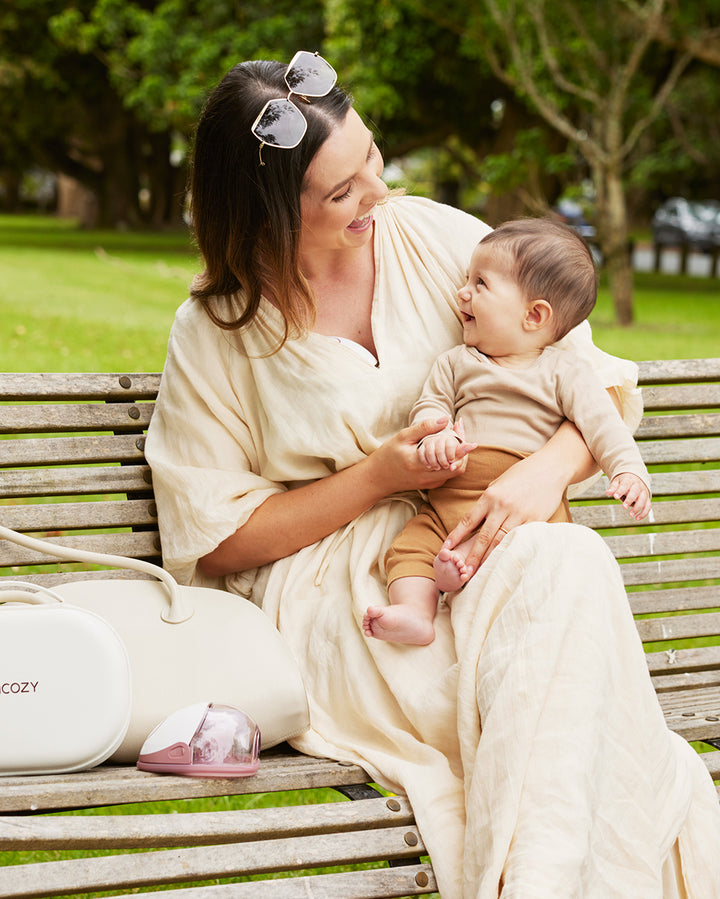 Momcozy M6 hands-free breast pump on bench next to smiling mother with baby in park.